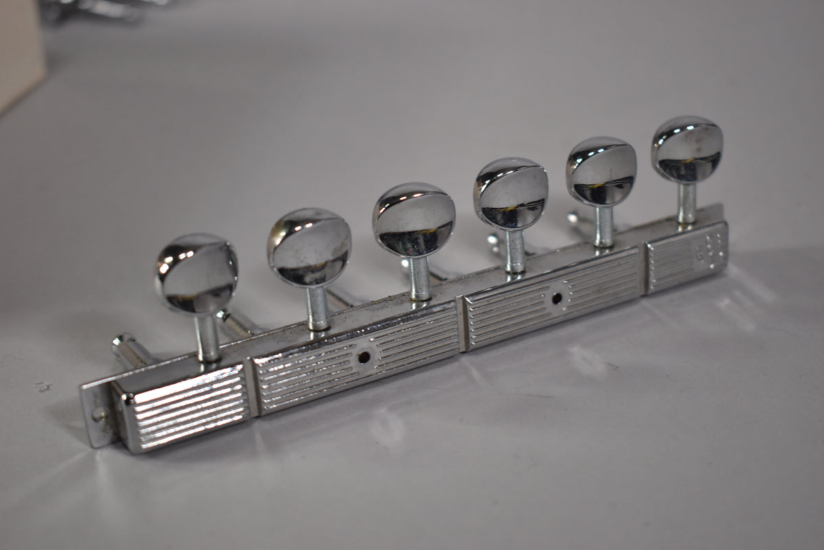 A set of six chrome tuners with oval buttons, mounted on a ribbed metal strip, viewed from above at an angle.
