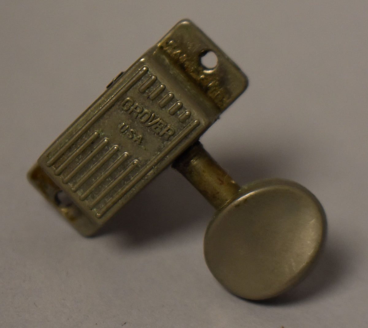 A single, tarnished nickel-plated tuner is angled, showing its ribbed casing, mounting screw hole, and round button with visible wear.
