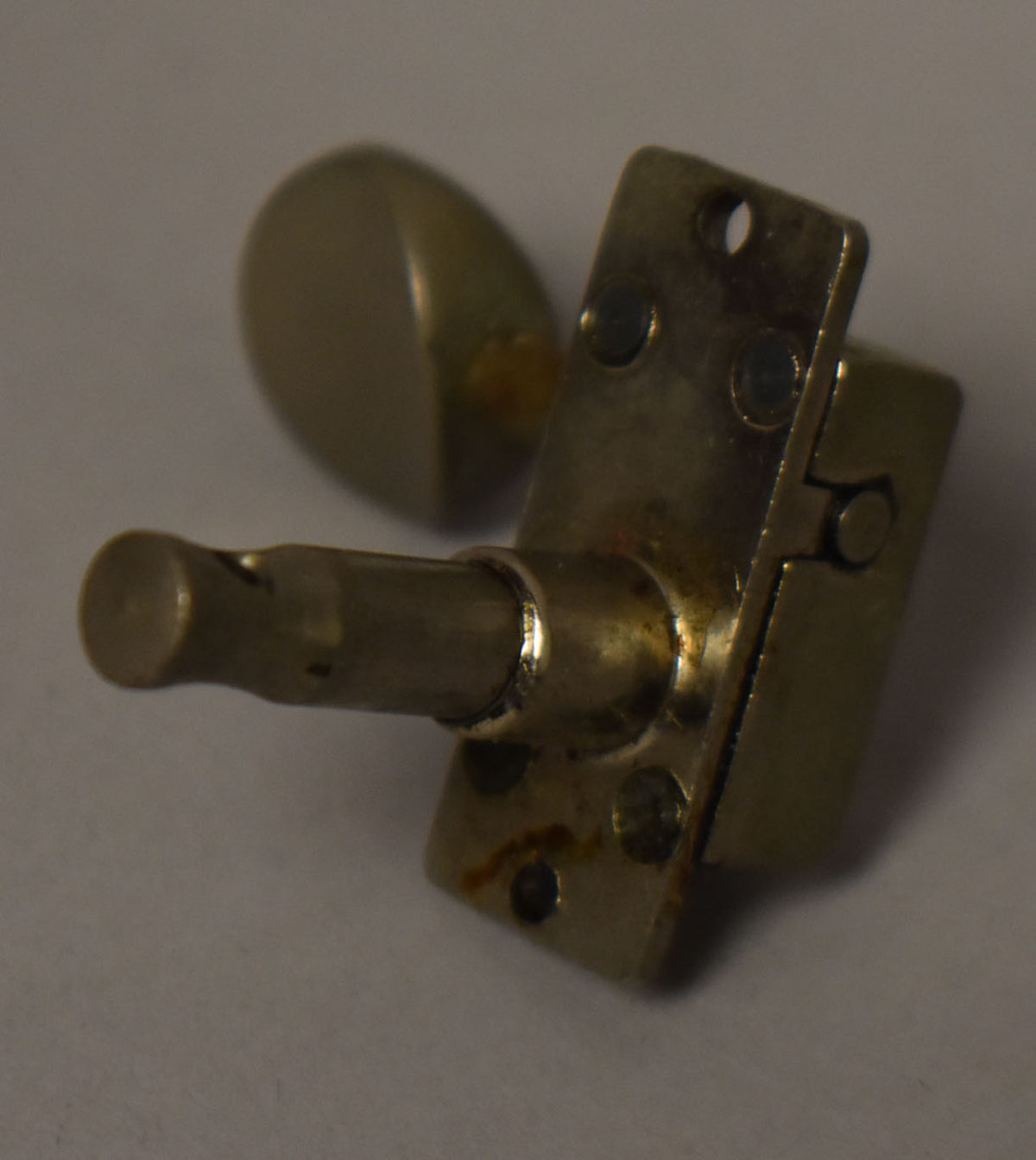 A single, tarnished gold-plated tuner is angled from above, showing the rectangular base with two screw holes and the tuning peg.