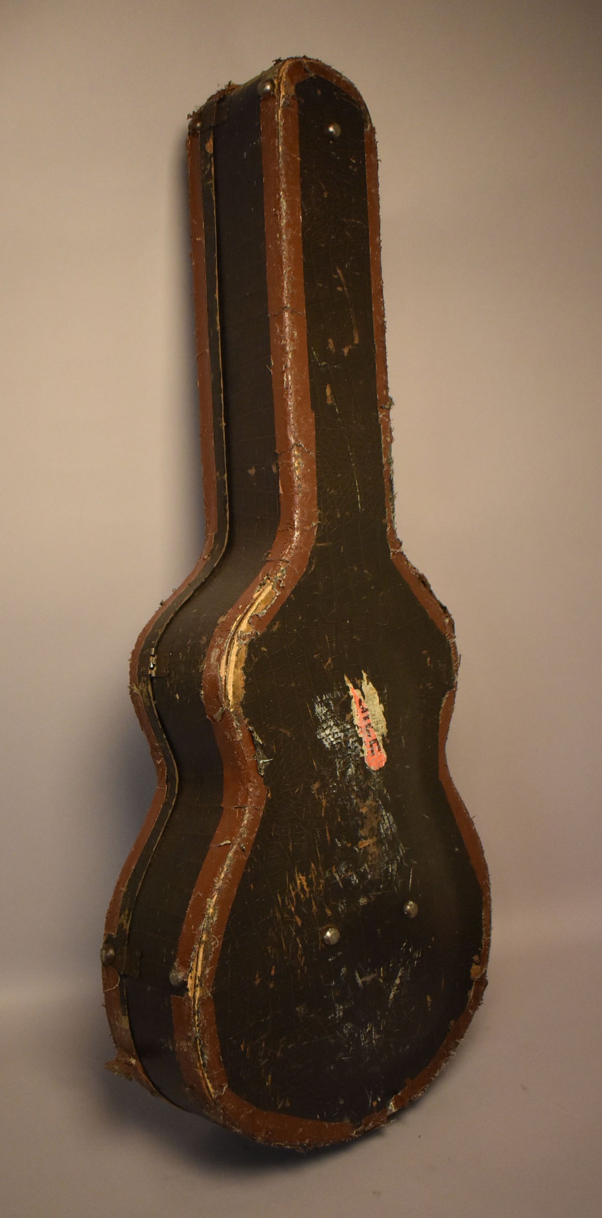 The guitar case stands upright, showing its dark, worn faux alligator finish with brown trim and exposed wood along the edges.