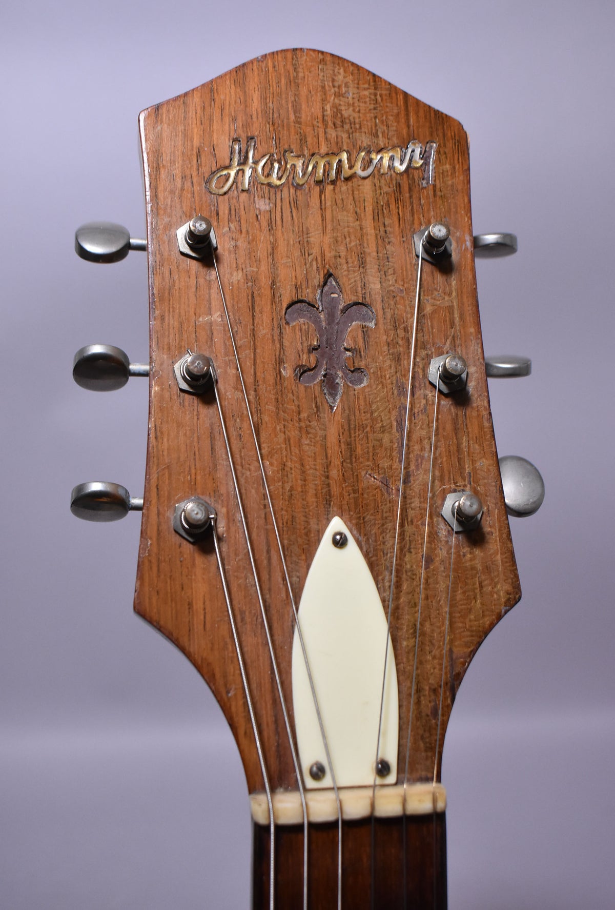 The guitar's natural wood headstock, with a carved fleur-de-lis, shows wear and features six metal tuners and a cream-colored truss rod cover.