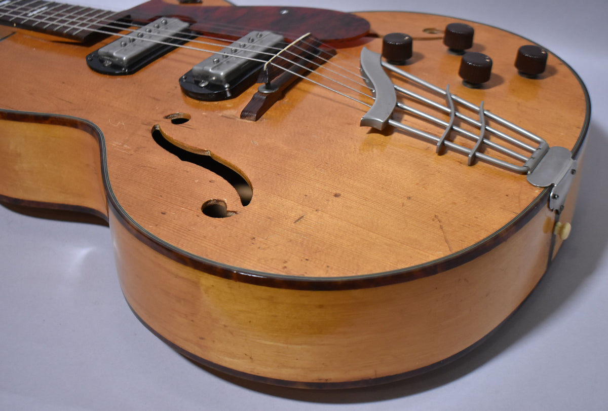 The lower bout of a natural finish archtop electric guitar, showing the f-hole, two pickups, a trapeze tailpiece, and four dark brown control knobs.