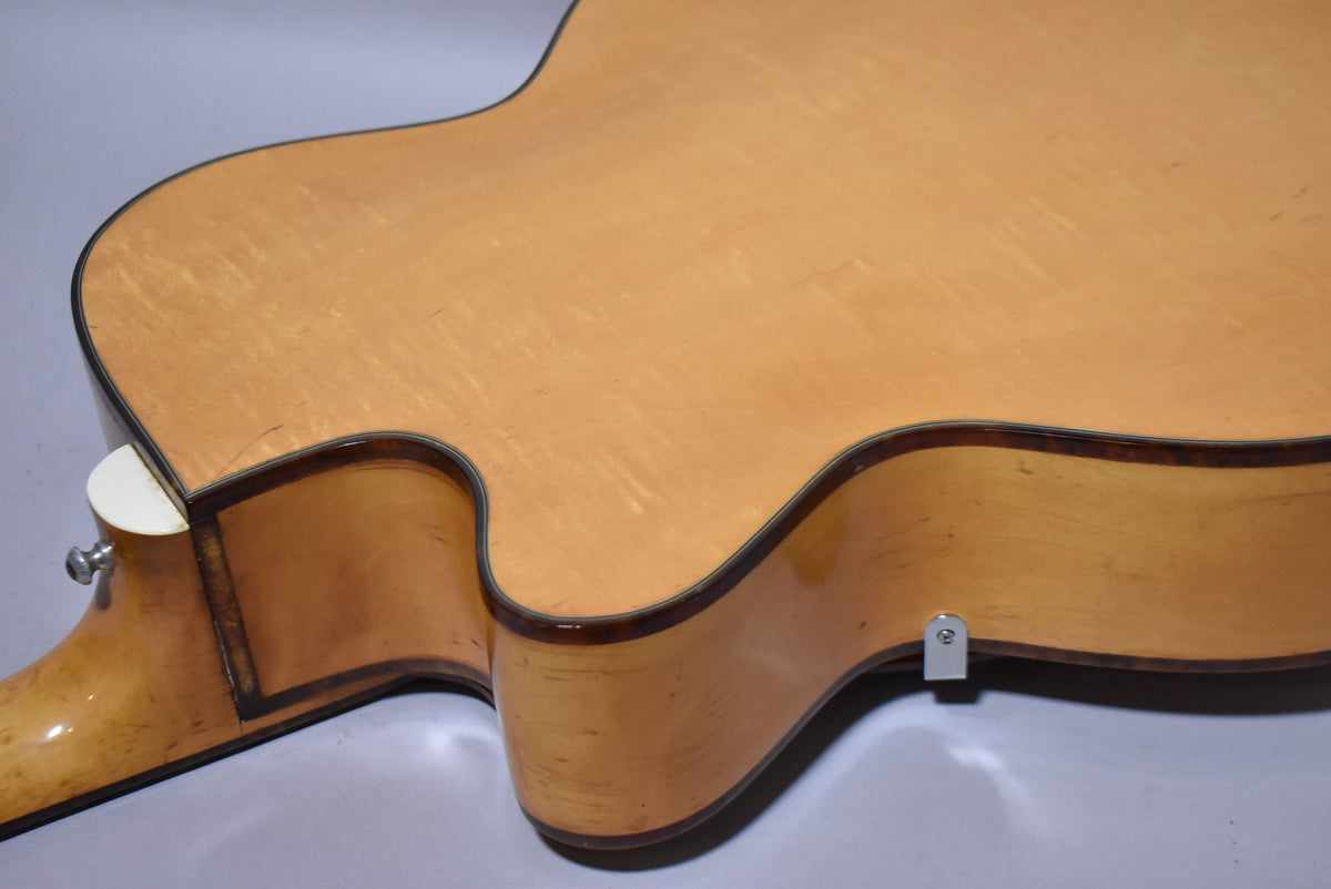 Close-up of the natural finish archtop guitar's lower bout and cutaway, showing binding, a strap button, and light checking.