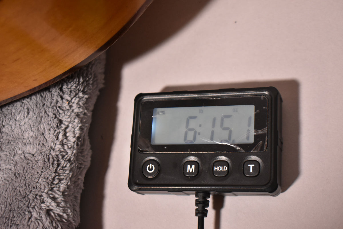 Close-up of the guitar's natural finish body resting on a grey cloth, with a digital scale displaying "6:15.1" in pounds and ounces.