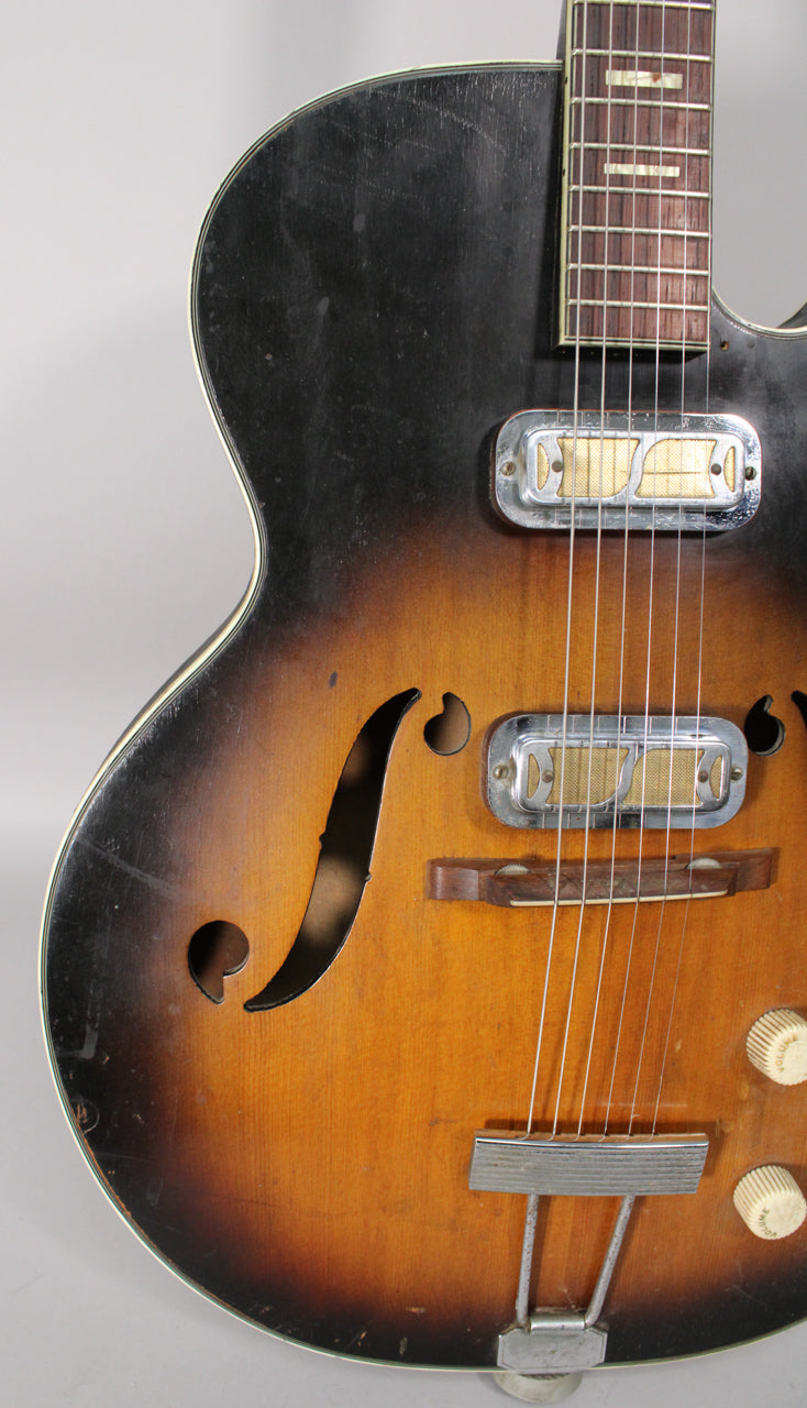 Close-up of a sunburst hollow body electric guitar, showing the lower bout, f-holes, two pickups, bridge, and two cream control knobs.