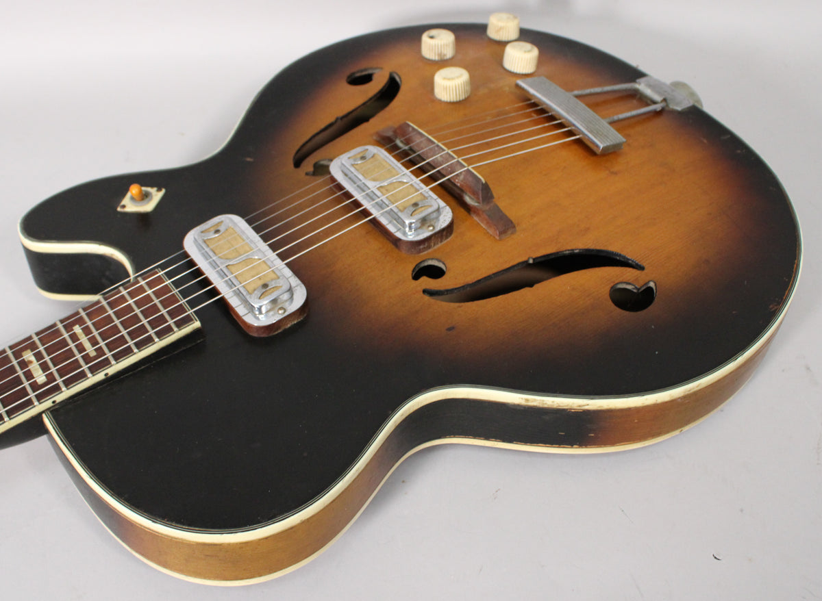 The guitar's sunburst body, showing wear, features two chrome-covered pickups, four cream control knobs, and a trapeze tailpiece.