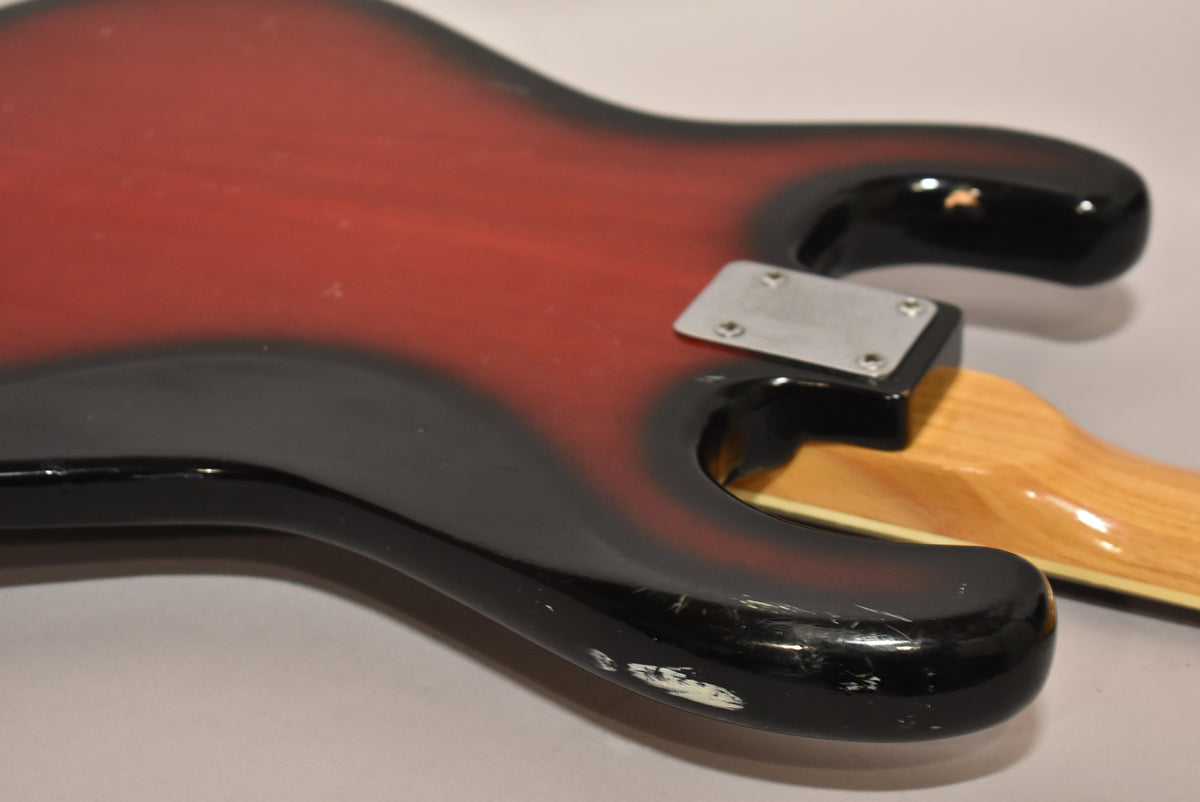 Close-up of the bass body's lower bout, showing the red burst finish, neck plate, and significant wear and chips on the black edge.