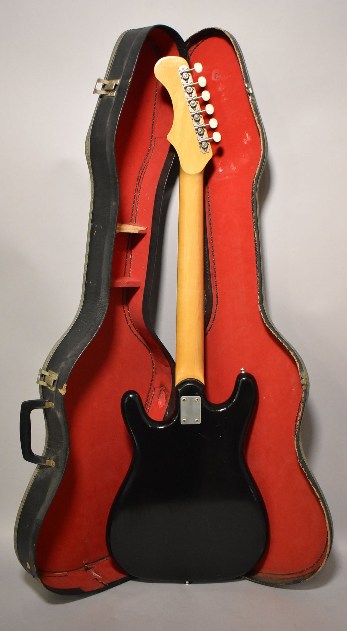 The black solidbody electric guitar is seen from the back, with its maple neck and headstock resting in a red-lined hard case.