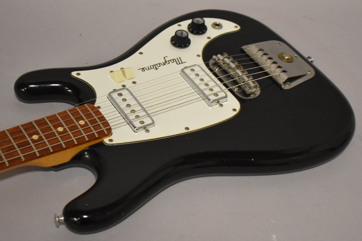 Black solidbody electric guitar, angled from above, showing the body, white pickguard, two single-coil pickups, and a vibrato tailpiece.