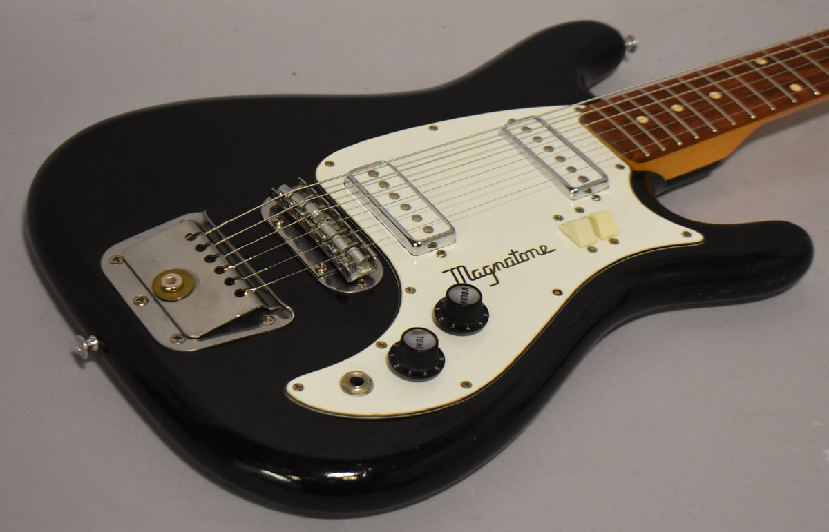 Black solidbody electric guitar, angled from the lower right, showing the body, white pickguard, two pickups, and two black control knobs.