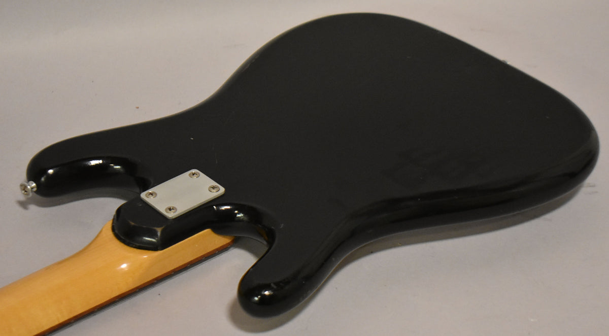The black solidbody guitar's back, angled from the lower bout, shows a light maple neck and a chrome neck plate with four screws.