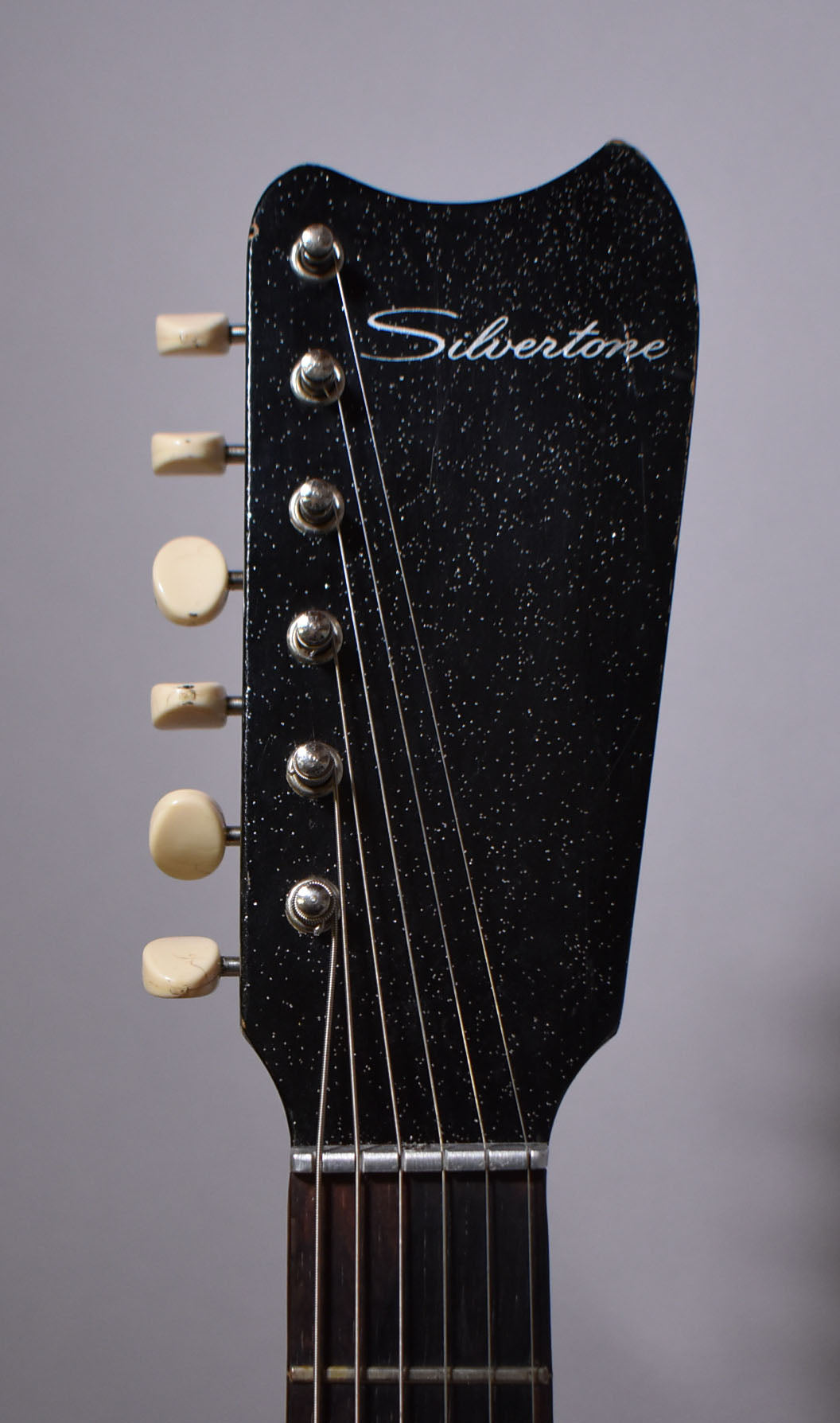 The guitar's headstock, angled slightly left, is black with silver sparkle, six cream tuning pegs, and light wear on the edges.