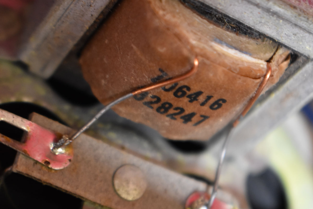 Extreme close-up of a pickup's copper coil, wrapped in aged brown paper with visible numbers, showing soldered wires and metal brackets.