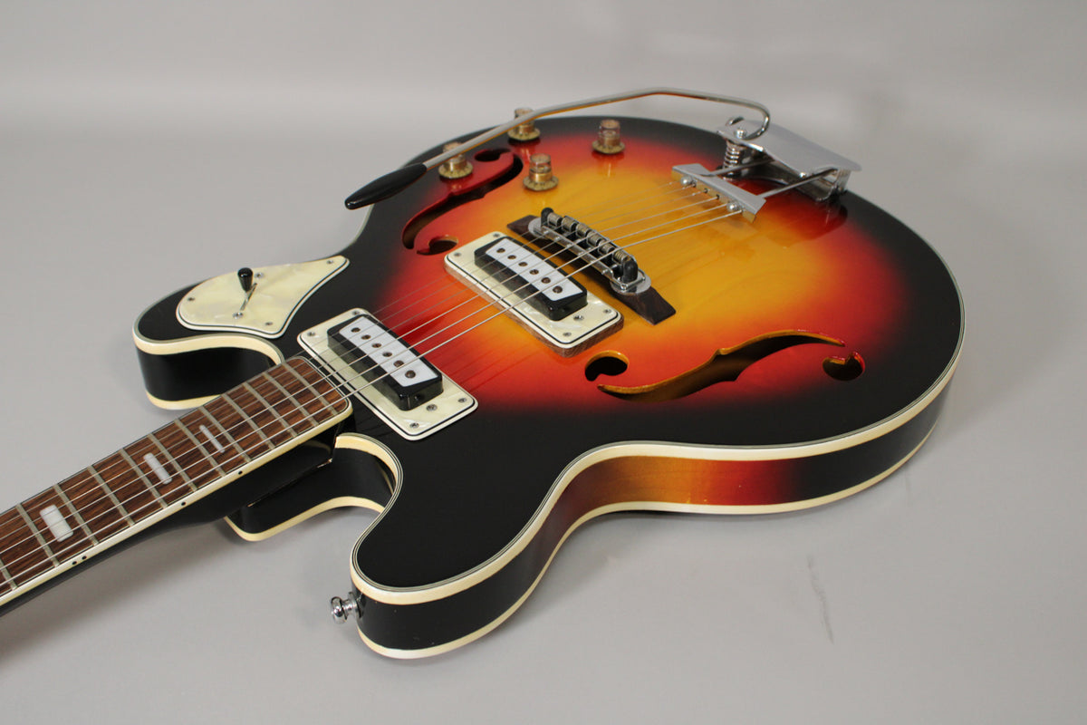 The sunburst hollowbody guitar is angled from the upper left, showing its two white-bound humbuckers, f-holes, and a Bigsby-style tremolo.