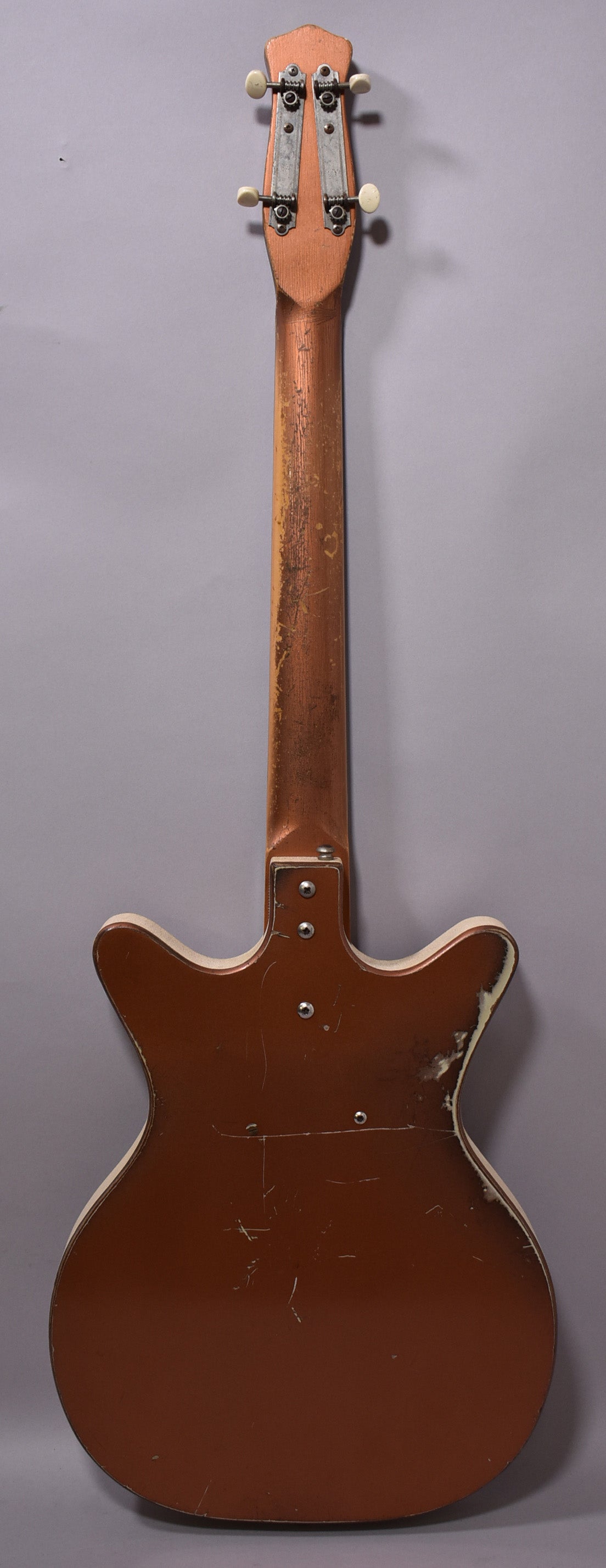 Full rear view of a copper-brown bass with a worn finish, showing a bolt-on neck, white binding, and four cream-button tuners.