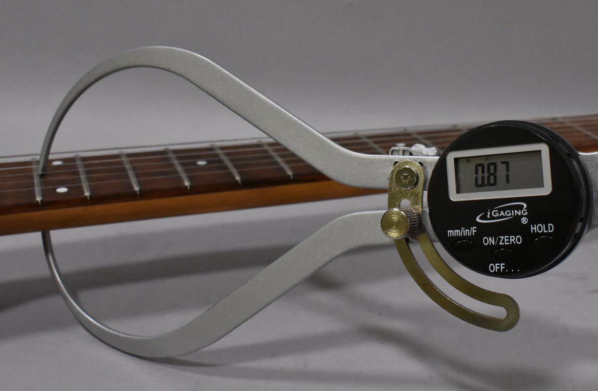 A digital caliper measures the fret height on the guitar's dark brown fretboard, showing "0.87" on its screen.
