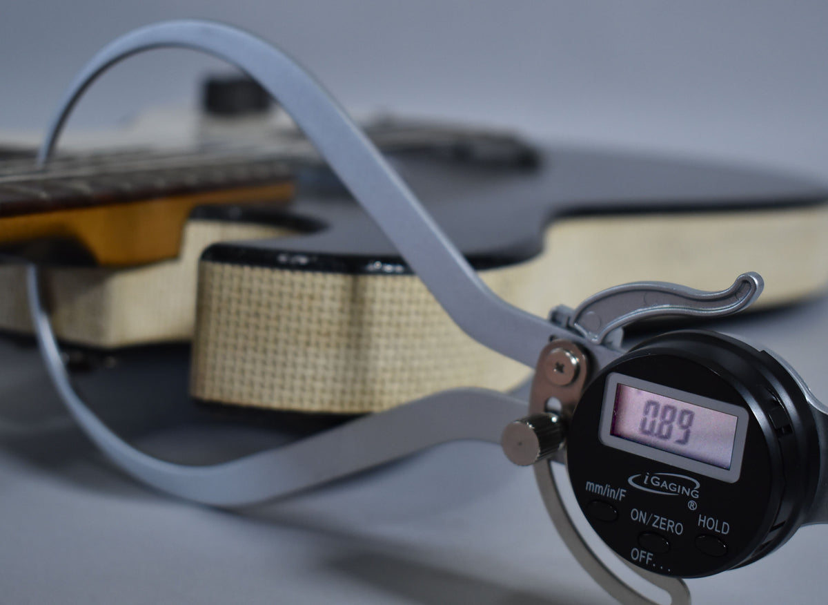A digital caliper measures the guitar's dark fretboard, showing "0.89" on its screen, with the black body and tan tweed case in the background.