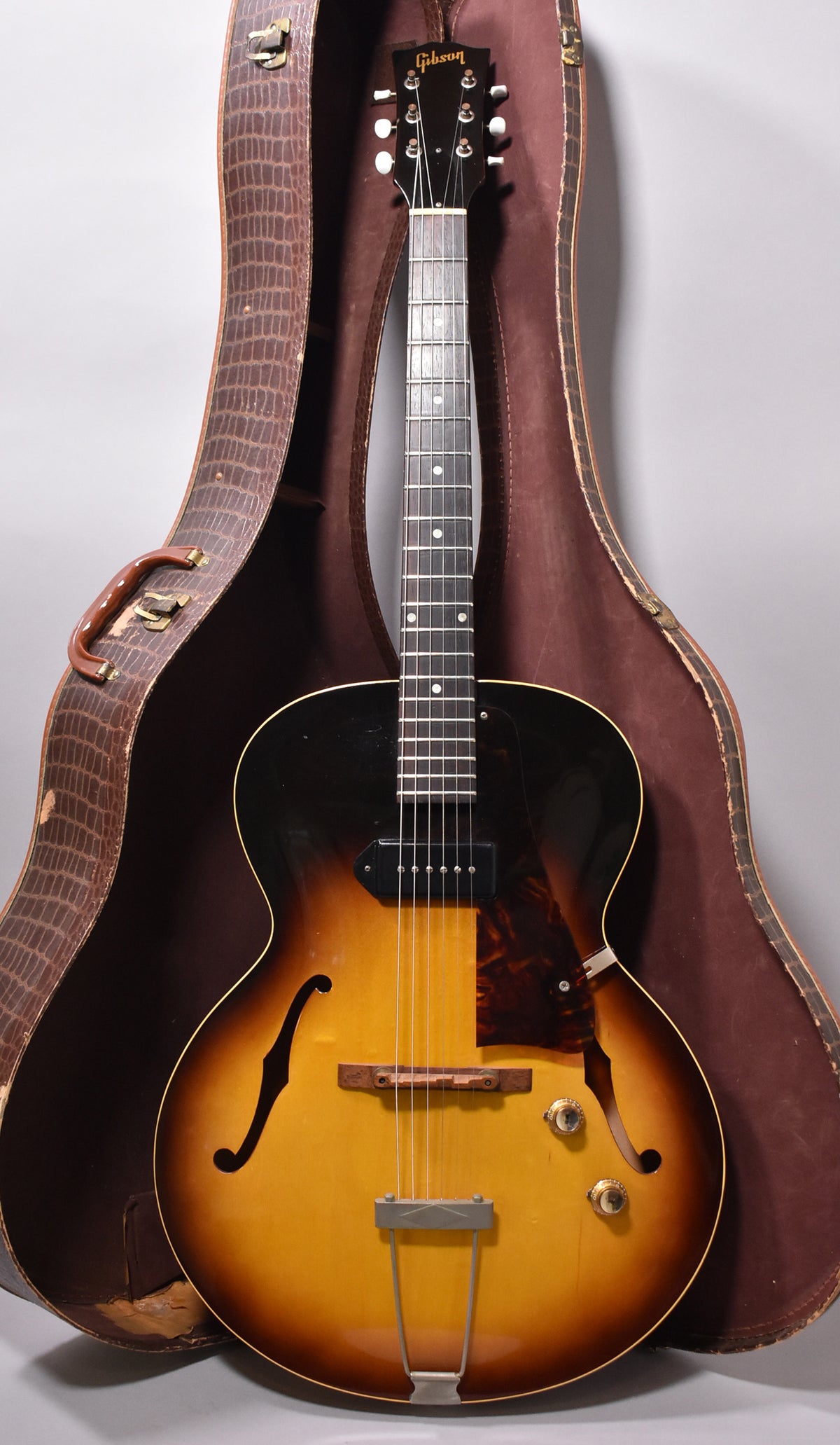 Full view of the sunburst archtop guitar in its open, brown alligator-patterned hard case, showing a single P90 pickup and two gold knobs.
