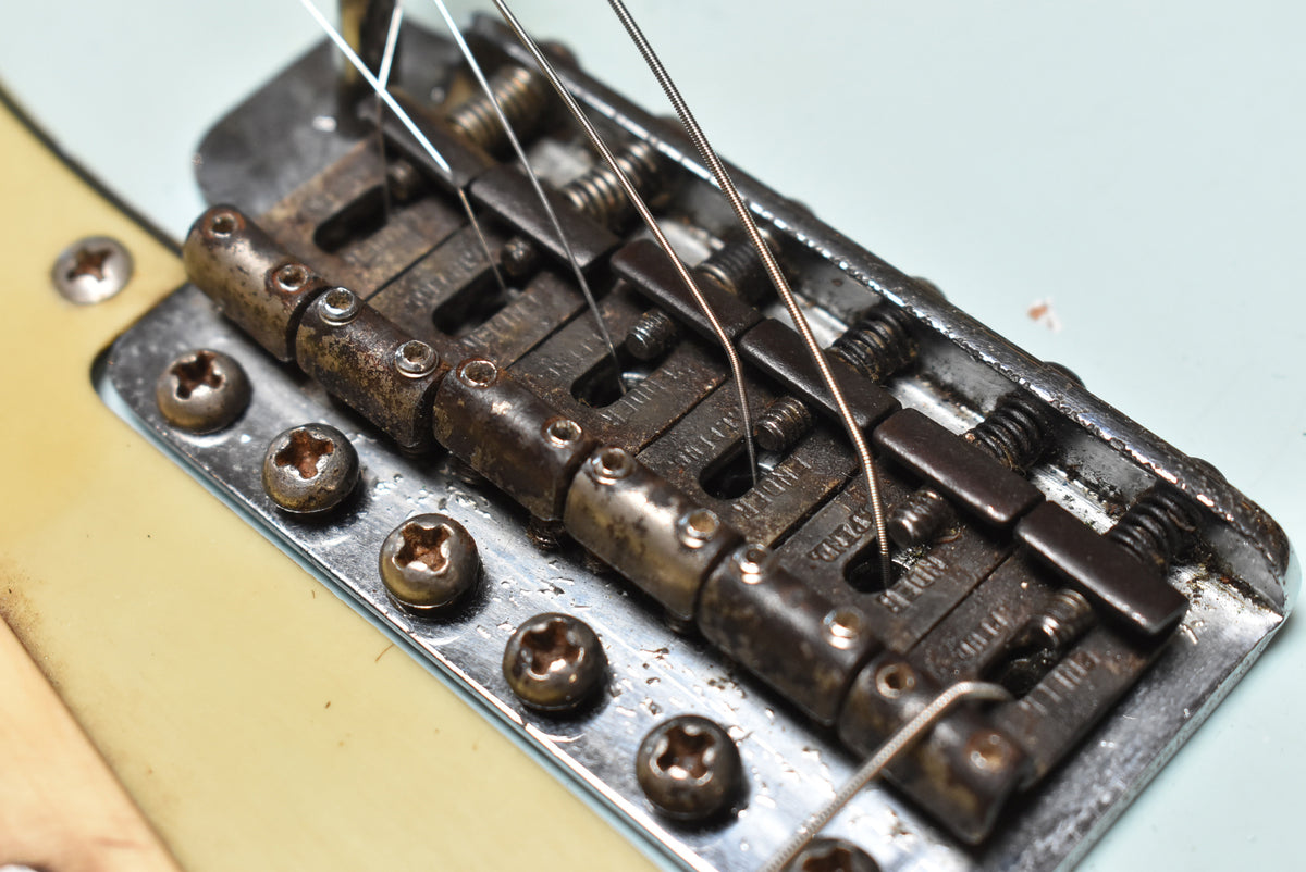 Close-up of the guitar's heavily rusted bridge and saddles, with six mounting screws and a light blue body visible.