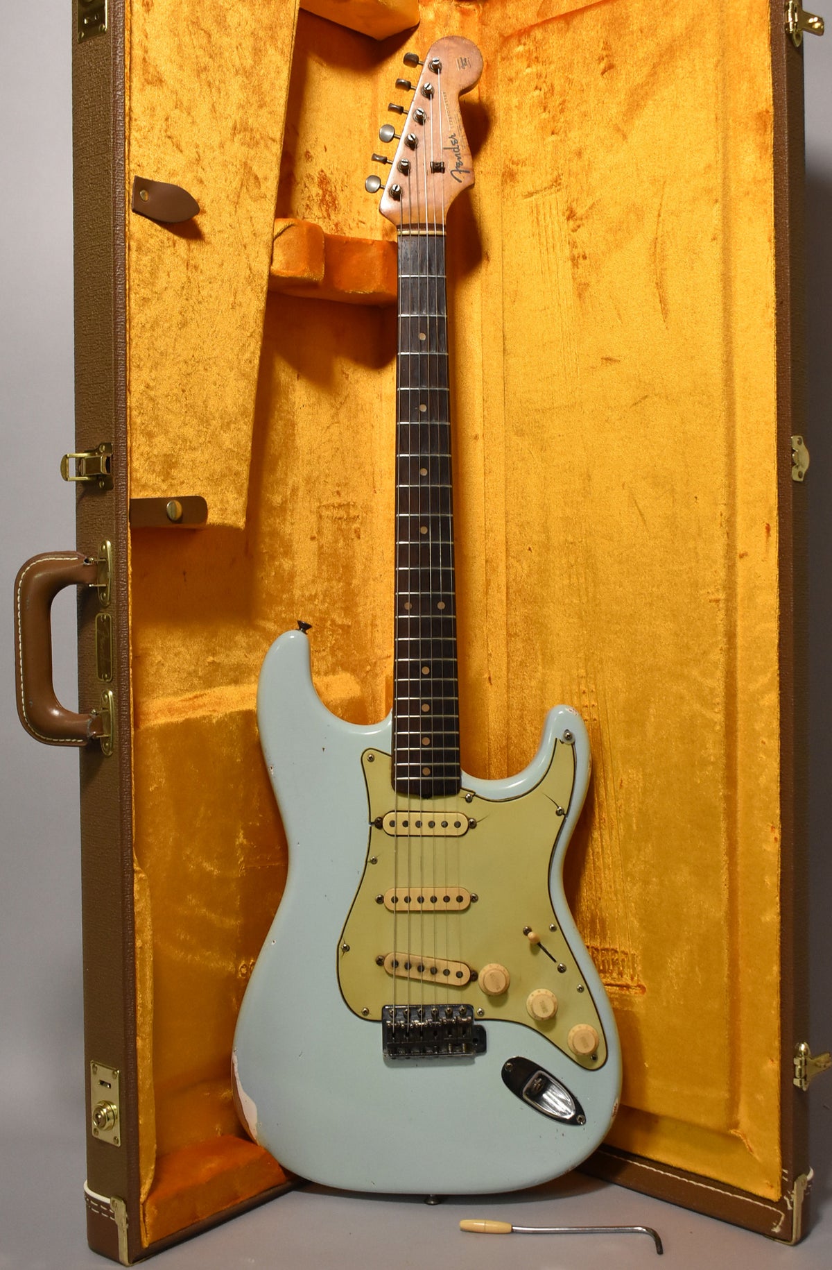 Full-length view of a light blue electric guitar with a cream pickguard, in a brown case with gold lining, showing wear on the body.
