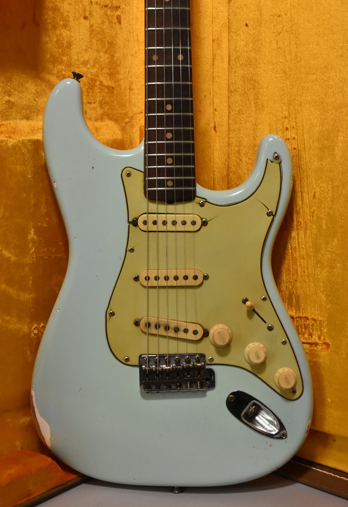 The aged sonic blue guitar, angled slightly right, shows a worn pale green pickguard, three cream pickups, and three volume/tone knobs.
