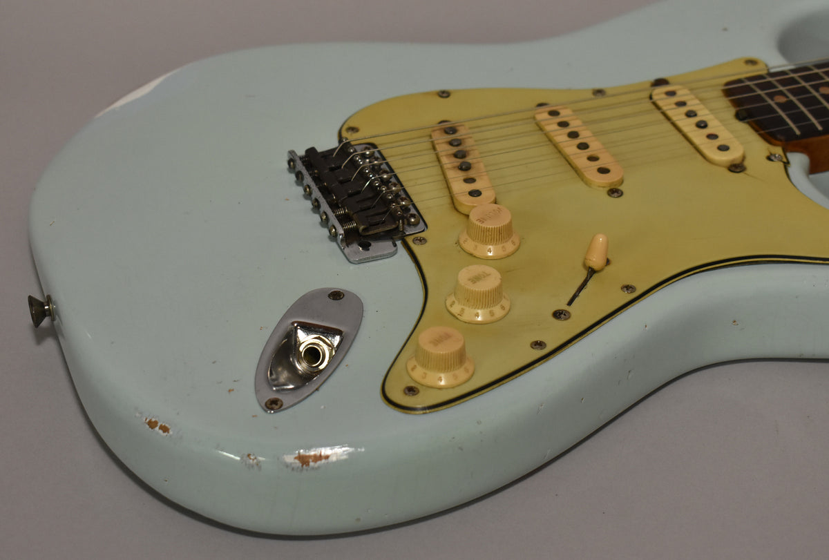 Close-up of the lower body of a light blue electric guitar, showing the worn output jack, bridge, aged cream pickguard, and controls.