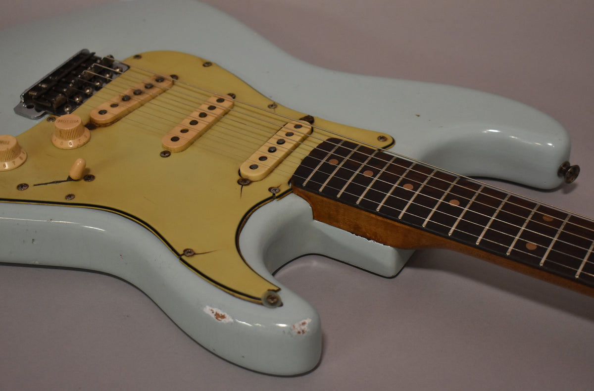 Light blue guitar body with a cream pickguard, three single-coil pickups, and two control knobs, showing paint chips and pickguard cracks.