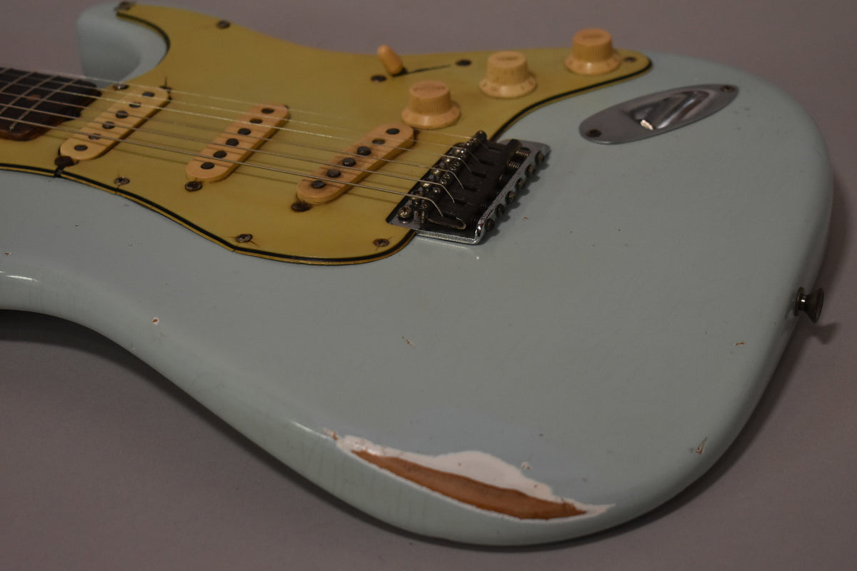 The aged sonic blue guitar body, with a large chip revealing wood, has a yellowed pickguard, three cream pickups, and four cream knobs.