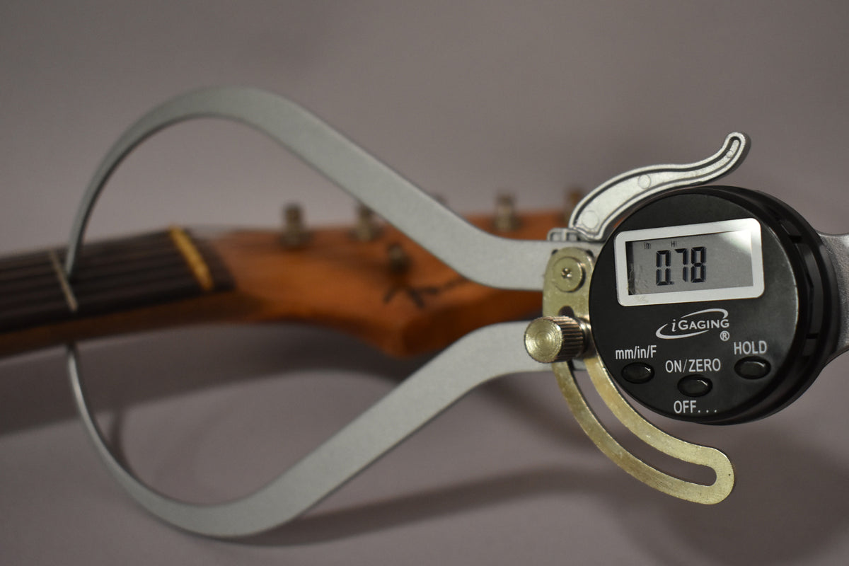 A digital caliper measures the guitar's neck at the first fret, displaying "0.78" against the worn, dark wood fretboard.