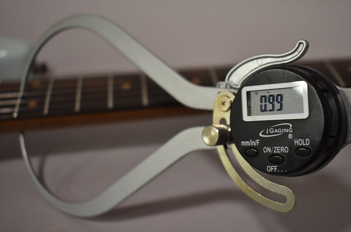 Close-up of a digital caliper measuring the guitar's dark rosewood fretboard, displaying "0.99" on its screen.