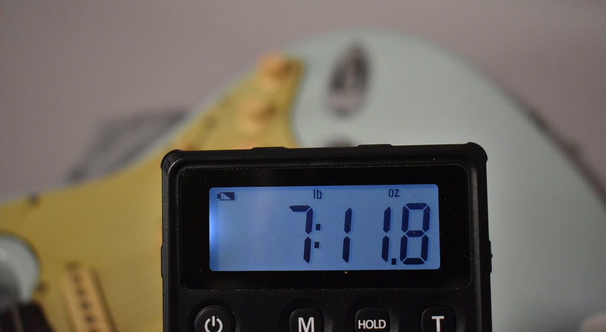 A digital scale displays "7:11.8 lb oz" in front of a light blue guitar body with a cream pickguard.