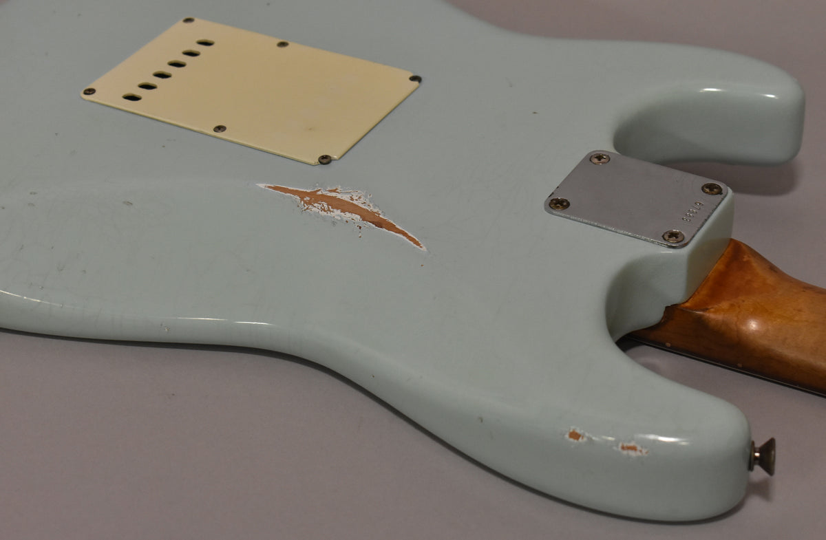 The back of a light blue guitar body, showing a large worn patch of exposed wood, a cream tremolo cover, and a metal neck plate.