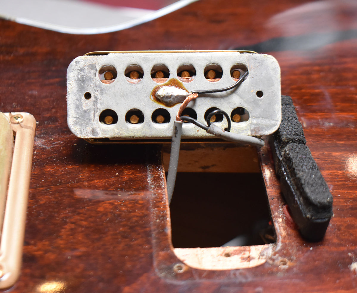 Close-up of a tarnished metal pickup with exposed wiring, removed from a dark walnut body, revealing the pickup cavity.