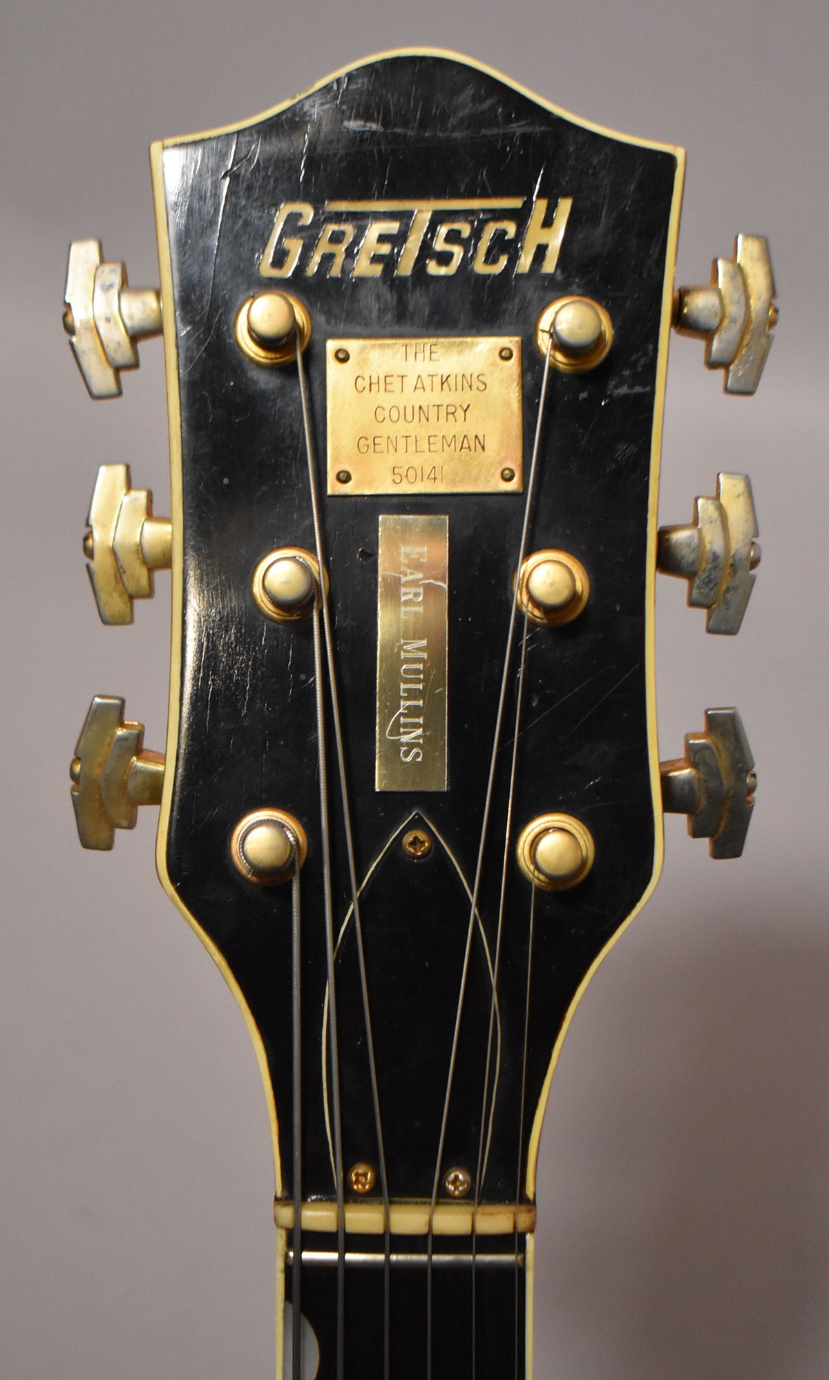 Close-up of the black headstock with gold tuners, showing finish checking and two brass plates, one engraved "Earl Mullins".