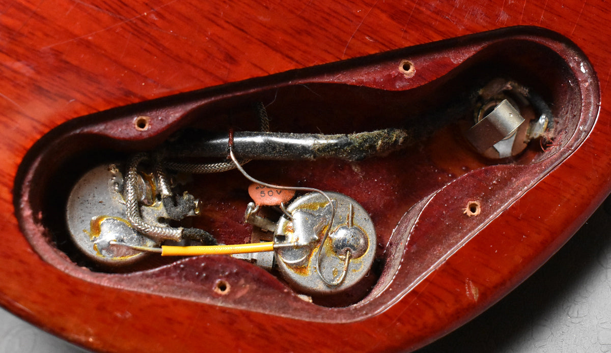 Close-up of the guitar's cherry red body, showing the control cavity with two tarnished pots, a 50V capacitor, and the output jack.