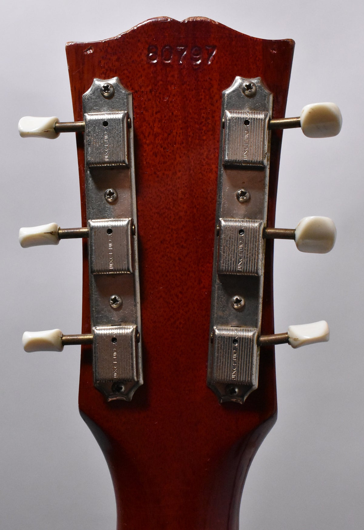The back of a cherry red headstock with six cream-buttoned tuners in two rows, showing light wear and a stamped serial number.