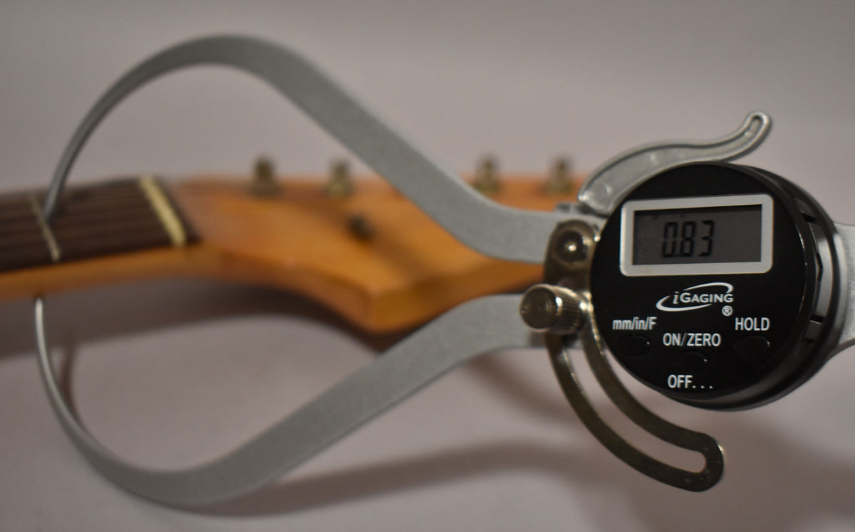 A digital caliper measures the guitar's neck, showing "0.83" on its screen, with the headstock and tuning pegs blurred in the background.
