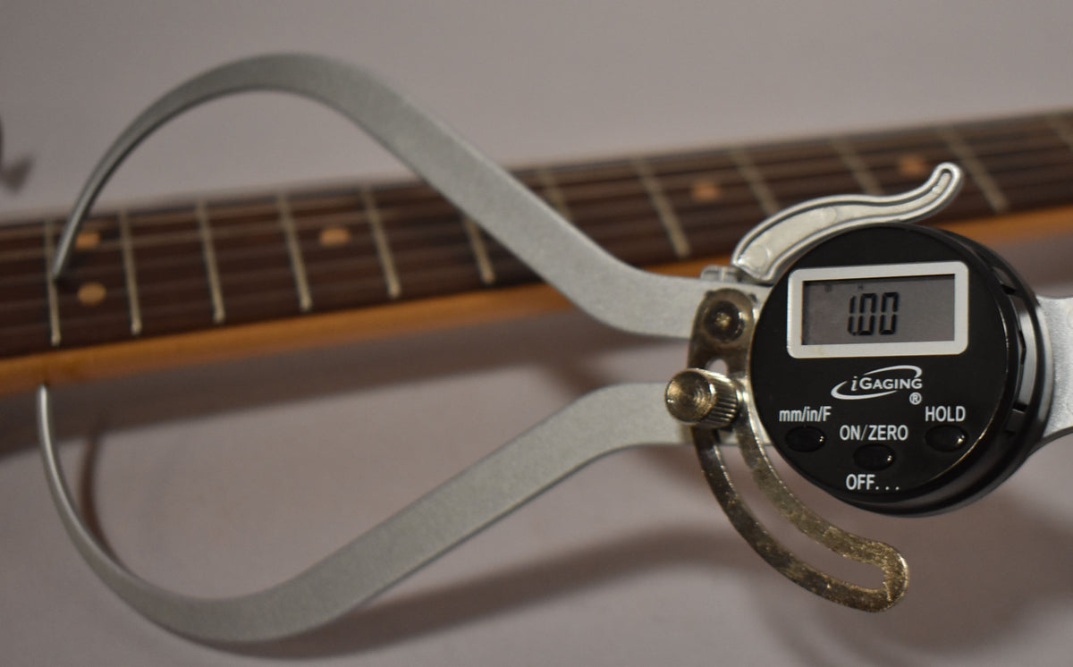 A digital caliper measures the guitar's fret height, showing "1.00" on its display, with the dark rosewood fretboard in the background.