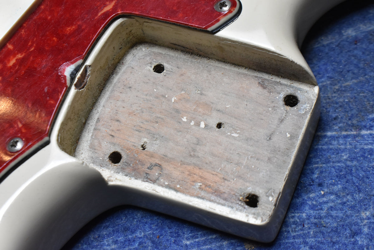 Close-up of the guitar's body cavity, showing exposed wood with screw holes, a red tortoiseshell pickguard, and chipped white finish.