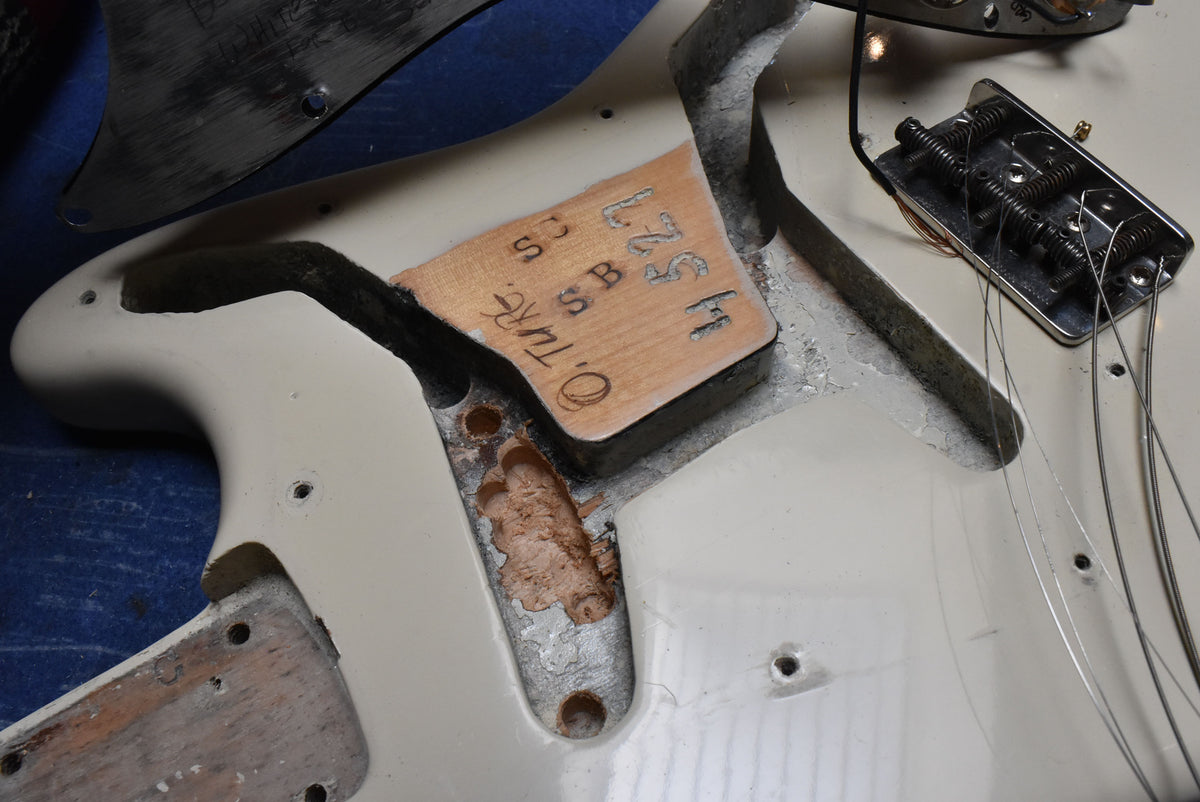 Close-up of the white guitar body's stripped wiring cavity, showing a wooden block with stamped numbers and a chrome bridge.