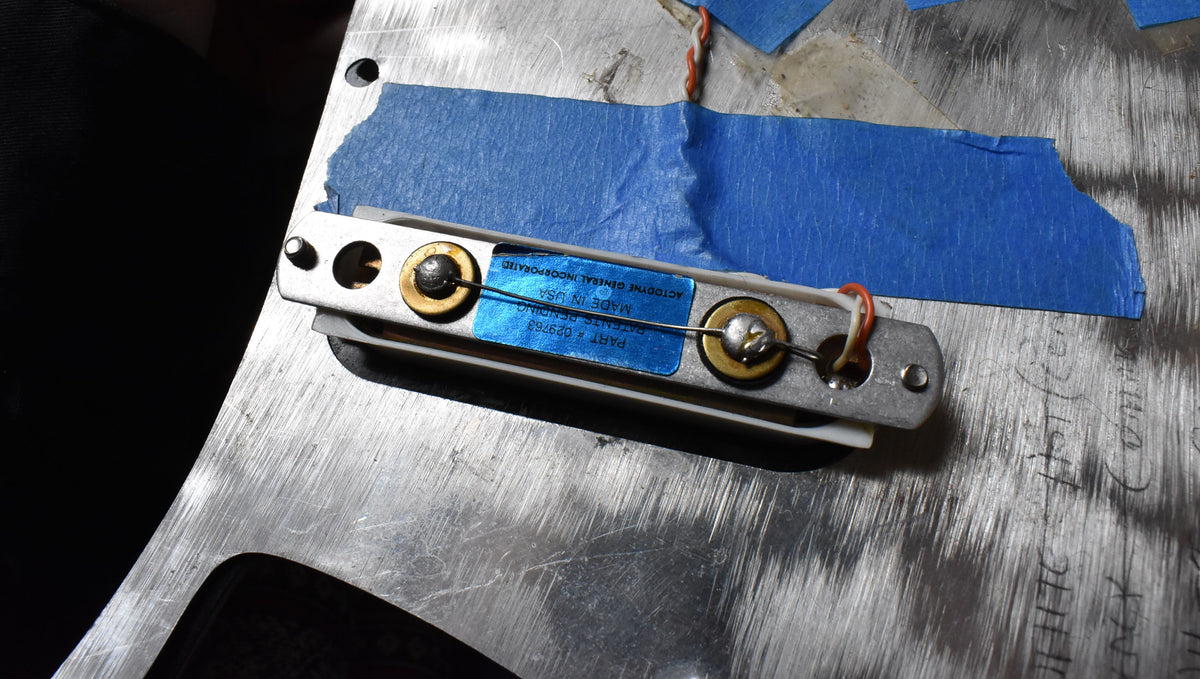 Close-up of a single-coil pickup with a blue label, mounted on a scratched metal plate with blue painter's tape.
