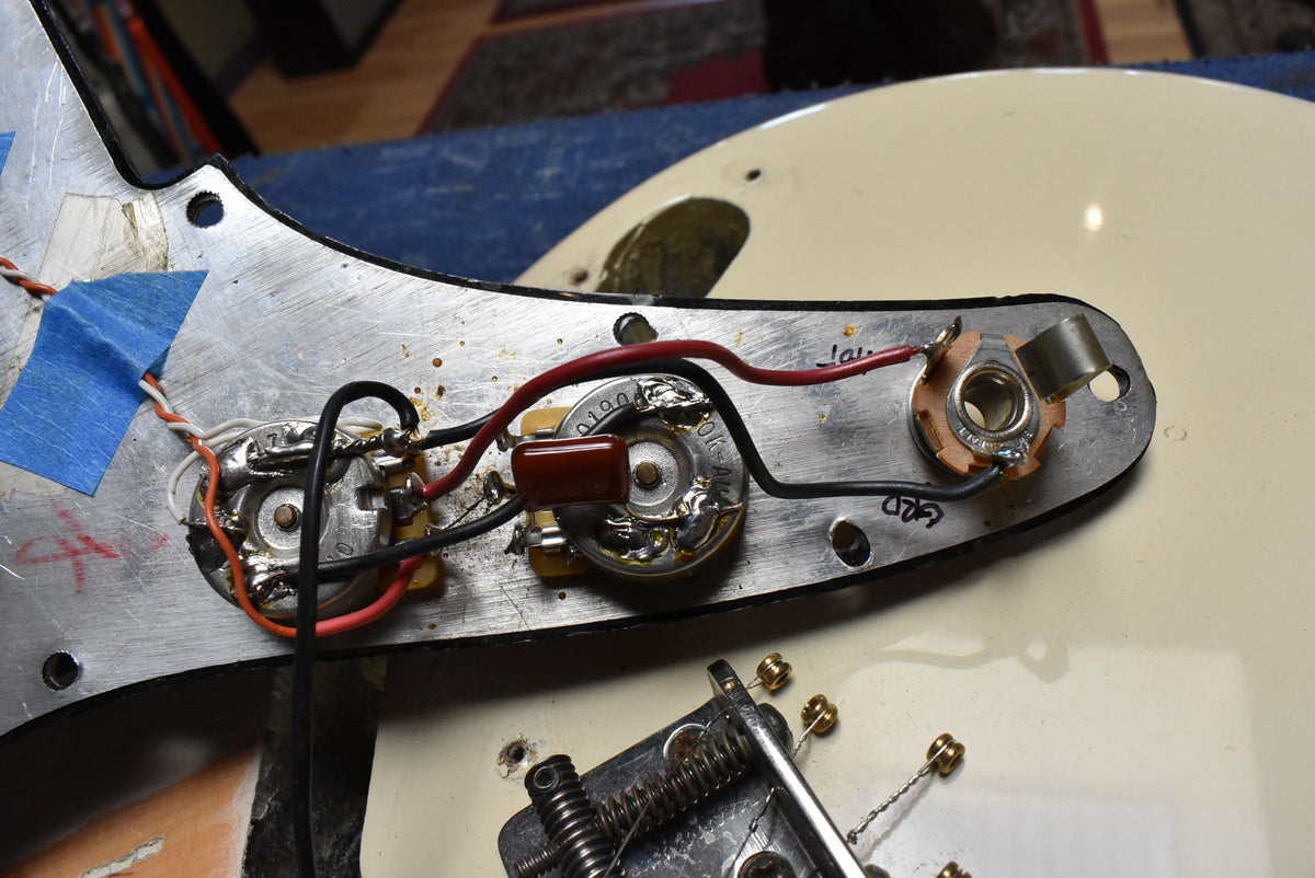 Close-up of the guitar's internal wiring, showing two pots, a capacitor, and the output jack, all soldered to a metal plate.