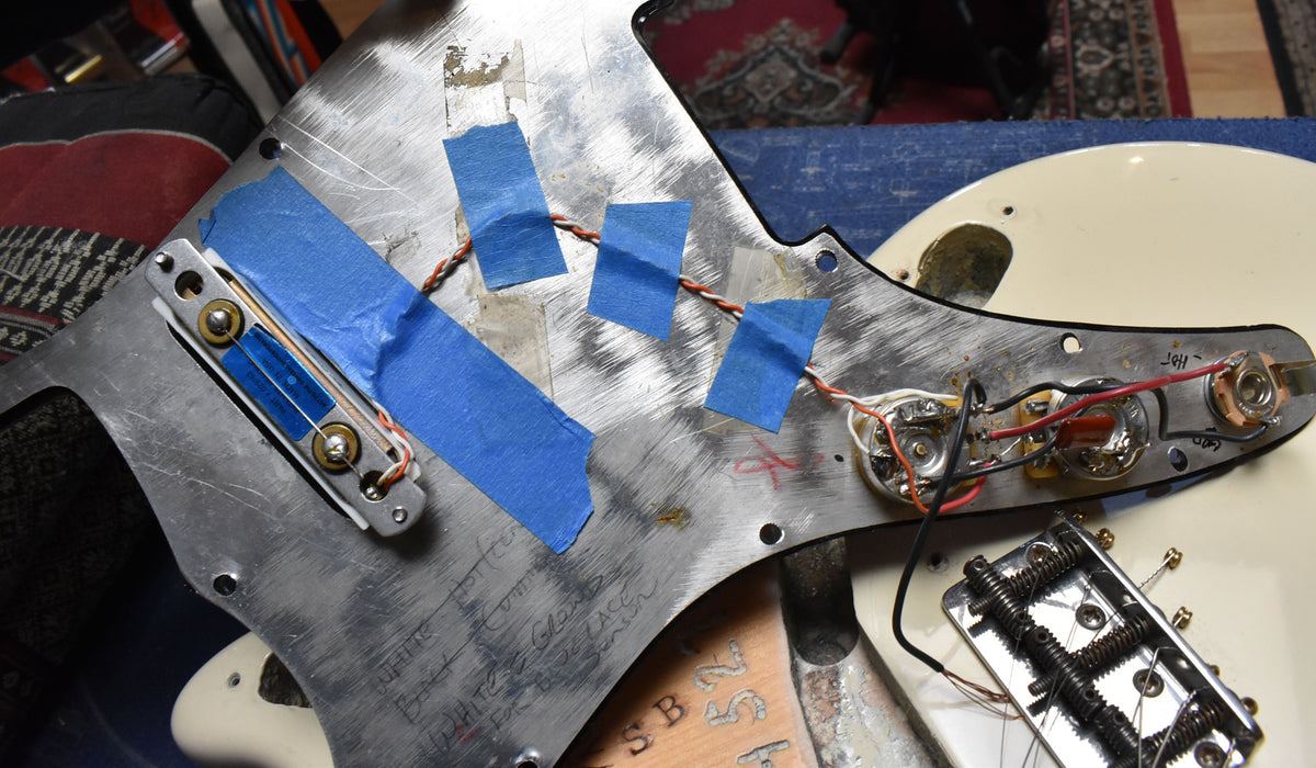 The guitar's body is visible with its pickguard removed, revealing the wiring, controls, and pickup, all secured with blue tape.
