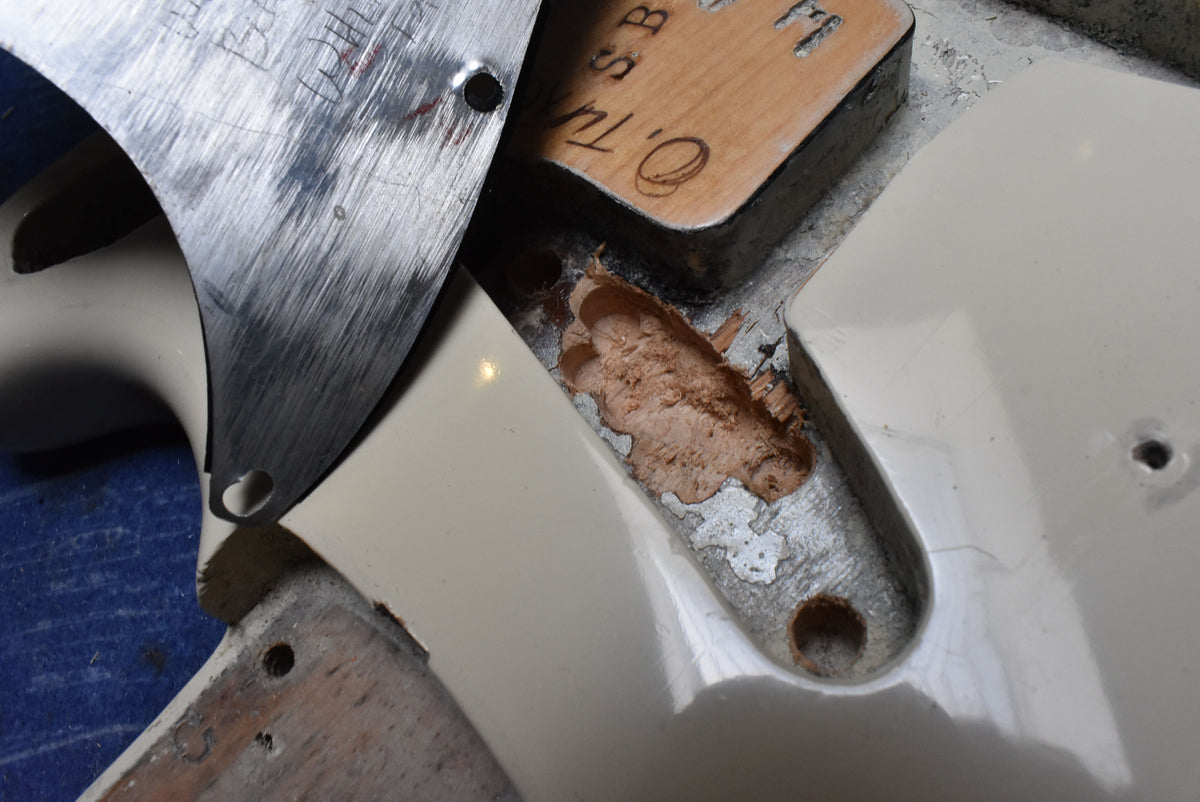Close-up of the guitar's body cavity, showing chipped white refinish, exposed wood, and a metal plate with handwritten markings.