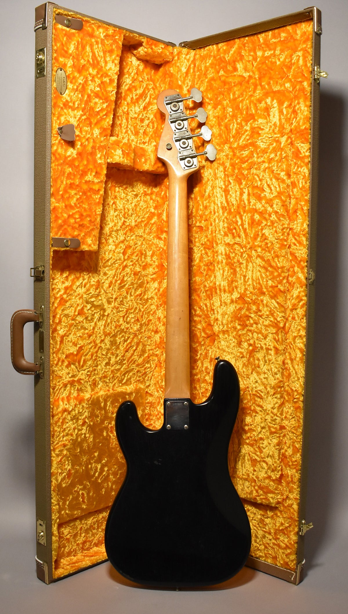 The black refinished bass is shown from behind, standing in its open, gold-lined hard case, with a worn maple neck and headstock.