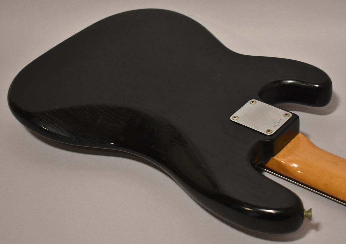The bass's black body, with visible finish wear and scratches, is seen from the back, showing the neck plate and a light wood neck.