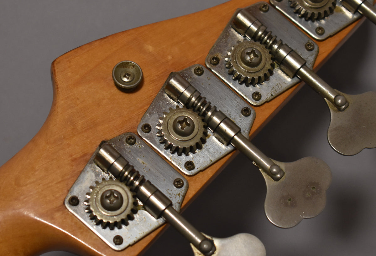 Close-up of the bass headstock, showing three worn, tarnished chrome tuners with exposed gears on a light wood finish.