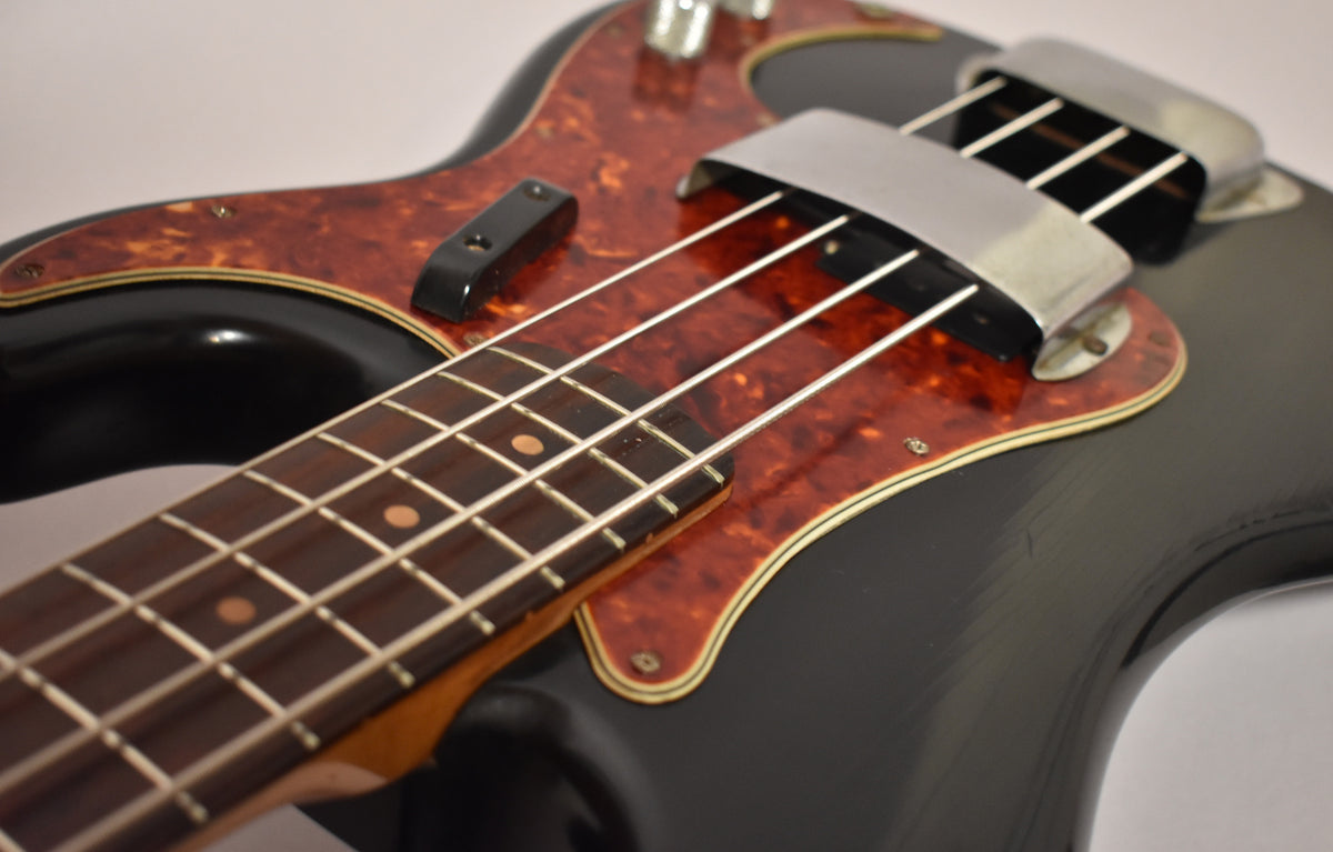 Close-up of a black bass body with a tortoiseshell pickguard, showing the lower fretboard, pickup cover, and some finish wear.