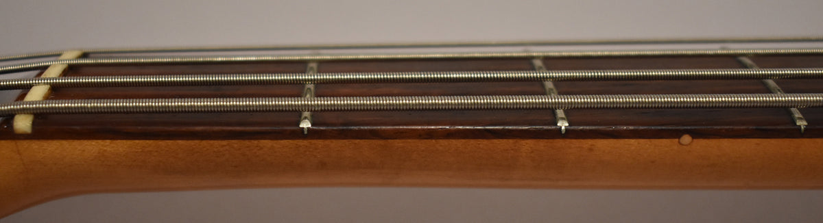 Close-up of the bass neck from the side, showing the dark fretboard, metal frets, four strings, and a small ding on the light wood neck.