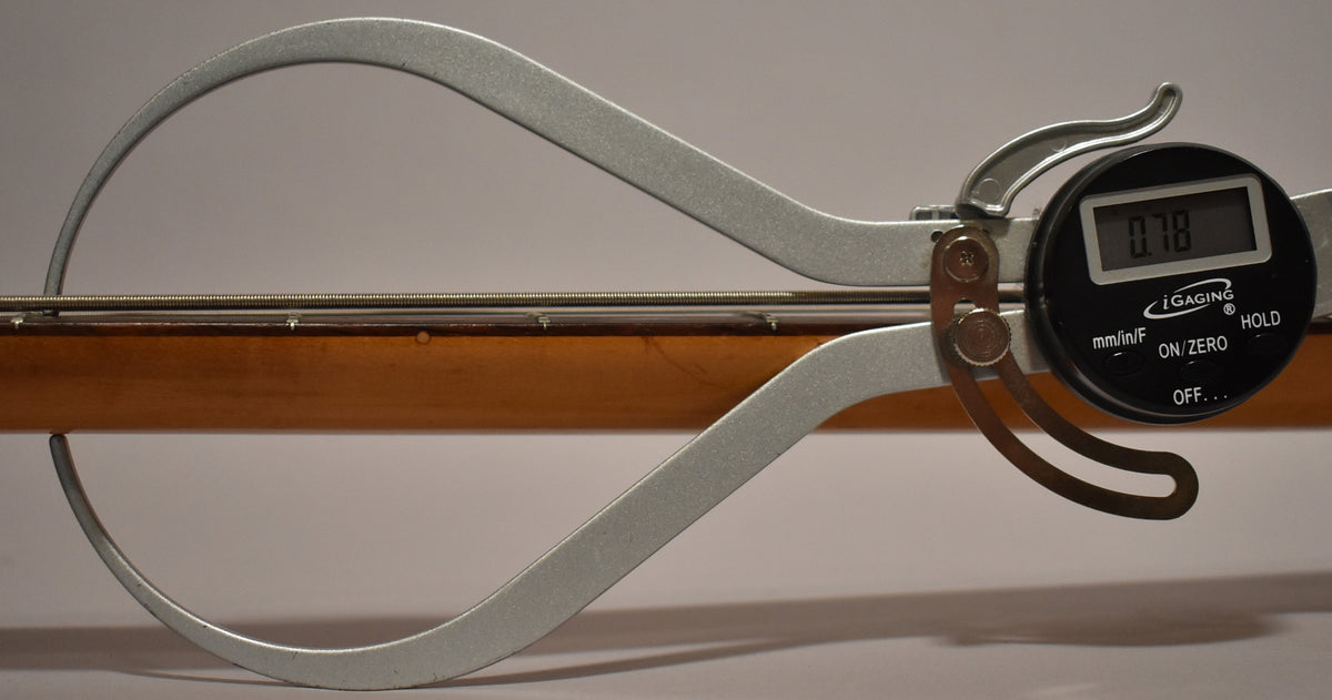 A digital caliper measures the neck relief of the bass, showing 0.78 on the display, with the strings and fretboard visible.